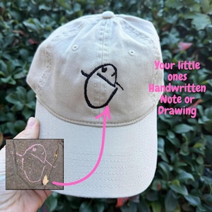 May include: A beige baseball cap with a black embroidered cartoon design. The cap features the text "Your little ones Handwritten Note or Drawing" in pink. A small drawing is shown in the lower left corner.