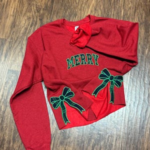 Pode incluir: Camisola vermelha com a palavra "MERRY" bordada na frente e laços de xadrez verde e preto nas mangas.