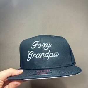 Puede incluir: Gorra de béisbol negra con visera plana y la inscripción bordada "Foxy Grandpa" en blanco. La gorra tiene una parte inferior blanca y un diseño estructurado, ideal para uso diario.
