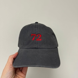 Può includere: Un cappellino da baseball grigio antracite con il numero "72" ricamato in rosso sul davanti. Il cappellino ha una visiera curva e una chiusura con cinturino in tessuto. Il cappello è realizzato in un materiale morbido e usurato.