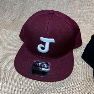 Puede incluir: Una gorra de béisbol de color granate con una letra J blanca en la parte delantera. La gorra tiene un cierre a presión y una etiqueta que dice "OTTO S-N-A-P ONE SIZE FITS MOST".