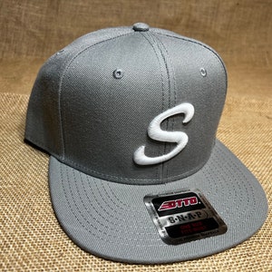 Puede incluir: Gorra snapback gris con una "S" blanca bordada en la parte delantera. La gorra tiene una visera plana y una corona estructurada. Una etiqueta en la visera dice "OTTO SNAP". Se muestra sobre una superficie texturizada de tonos neutros.
