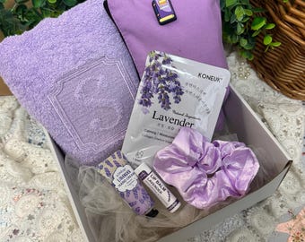 May include: A gift box filled with lavender-scented self-care items. Includes a lavender towel, pillow, face mask, hand cream, lip balm, and a scrunchie. The products are arranged in a white box, with a sprig of greenery in the background.