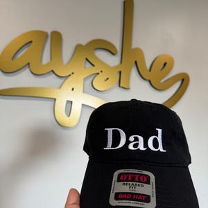 Peut inclure: Casquette noire avec le mot "Dad" brodé en blanc. La casquette a une coupe décontractée et une visière incurvée. L'étiquette indique "OTTO RELAXED FIT DAD HAT MAXIMUM COMFORT".