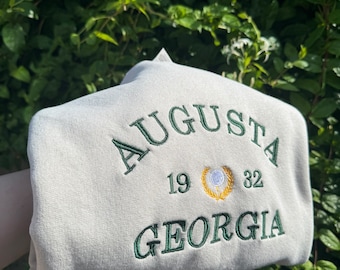 May include: A light gray sweatshirt with green embroidered text that reads "AUGUSTA 19 32 GEORGIA". A green and gold embroidered golf ball logo is between the numbers.