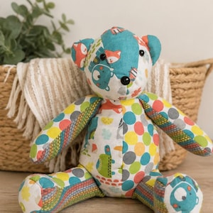 May include: A stuffed bear toy constructed from patchwork fabric featuring a variety of colourful patterns. The bear has black button eyes and a black button nose, with limbs and a torso decorated in polka dots, animal illustrations, and geometric prints.