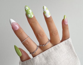 May include: Close-up of a hand with green and white nail polish. The nails are long and almond shaped. The green nails have a white floral design. The white nails have a light pink base.