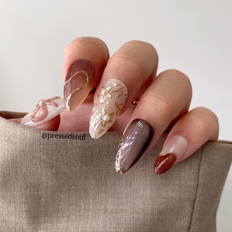 May include: Almond shaped nails with a brown ombre design, a white nail with gold floral design, and a brown nail with a shimmering effect. The nails are decorated with gold accents and small pearl-like embellishments. The text '@pressedsoul' is visible in the image.