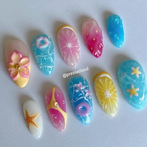 Pu&ograve; includere: Una collezione di dieci unghie acriliche con disegni a tema estivo. Le unghie presentano vari disegni, tra cui un fiore, una stella marina, un salvagente e fette di frutta. I colori includono rosa, giallo, blu e bianco. Le unghie sono disposte su una superficie bianca.