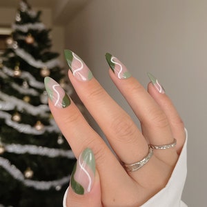 Op de afbeelding: Close-up van een hand met groene en witte abstracte nagelkunst. De nagels zijn lang en amandelvormig. De hand draagt een zilveren ring.