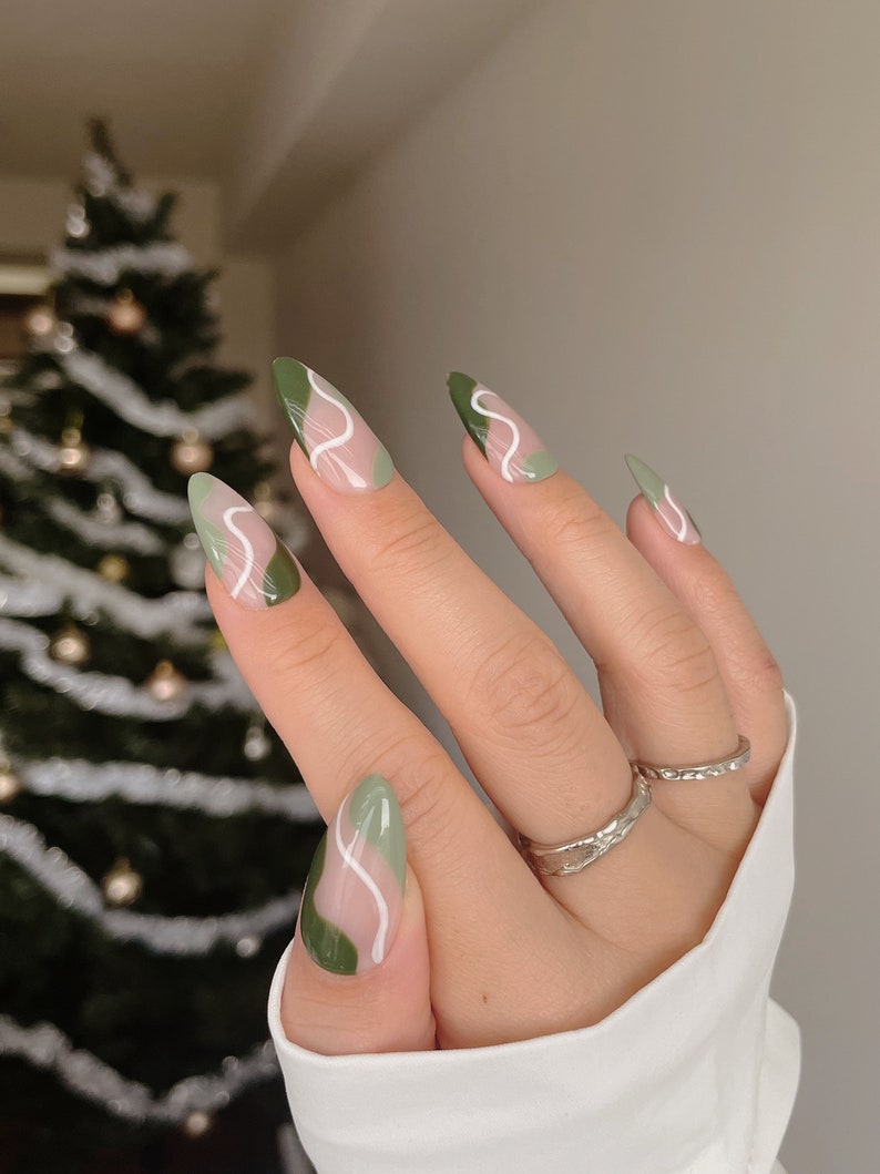 Op de afbeelding: Close-up van een hand met groene en witte abstracte nagelkunst. De nagels zijn lang en amandelvormig. De hand draagt een zilveren ring.