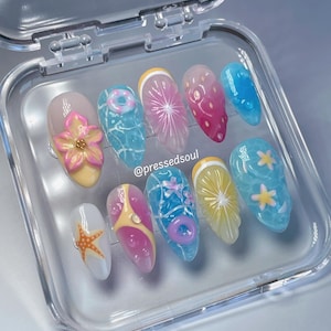 Pu&ograve; includere: Un set di dieci unghie finte con disegni a tema estivo. Le unghie presentano vari disegni, tra cui un fiore, una stella marina e fette di frutta. Le unghie sono esposte in una custodia di plastica trasparente.