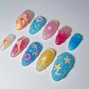 Pu&ograve; includere: Una collezione di dieci unghie finte con vari disegni. Le unghie presentano una gamma di colori, tra cui rosa, blu, giallo e bianco. I disegni includono fiori, frutta ed elementi a tema acquatico. Le unghie sono disposte su una superficie bianca.