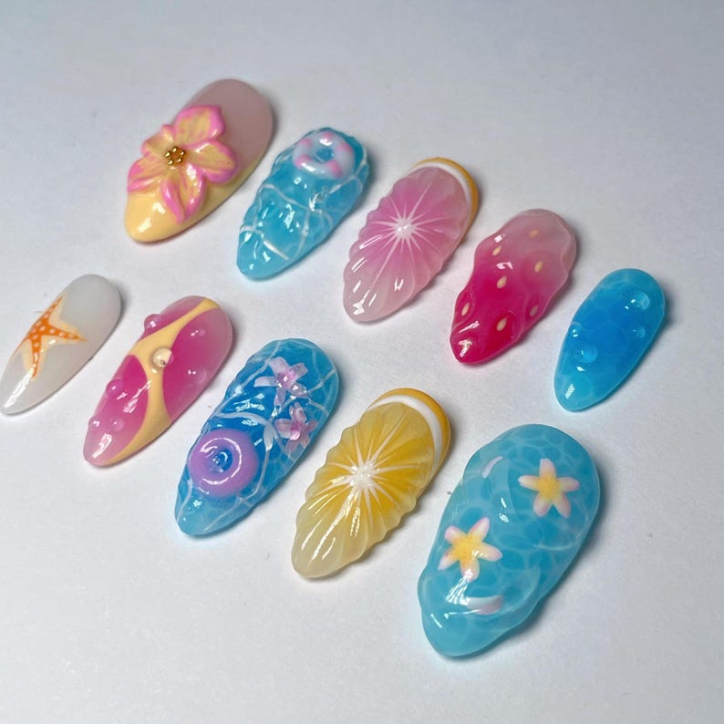 Pu&ograve; includere: Una collezione di dieci unghie finte con vari disegni. Le unghie presentano una gamma di colori, tra cui rosa, blu, giallo e bianco. I disegni includono fiori, frutta ed elementi a tema acquatico. Le unghie sono disposte su una superficie bianca.