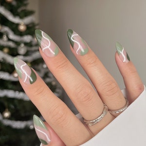 Op de afbeelding: Close-up van een hand met lange, amandelvormige nagels die zijn gelakt met een abstract ontwerp in groen en wit. De nagels zijn gelakt met een lichtgroene basis en hebben witte lijnen die een golvend patroon cre&euml;ren.