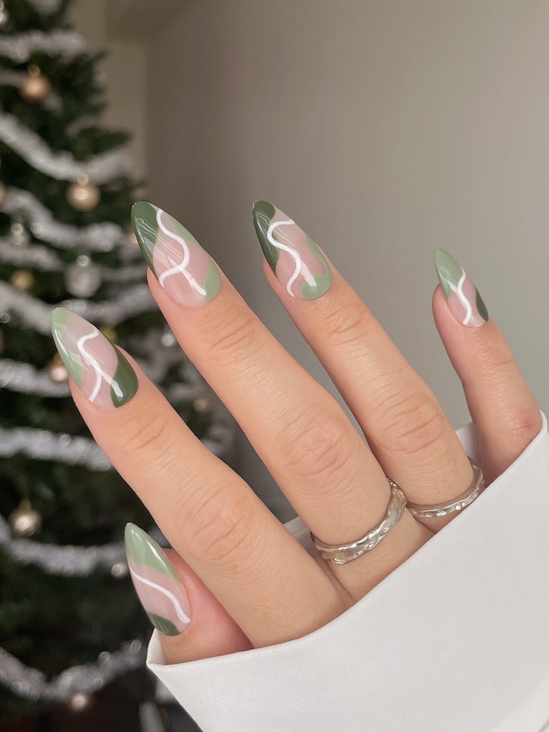 Op de afbeelding: Close-up van een hand met lange, amandelvormige nagels die zijn gelakt met een abstract ontwerp in groen en wit. De nagels zijn gelakt met een lichtgroene basis en hebben witte lijnen die een golvend patroon cre&euml;ren.