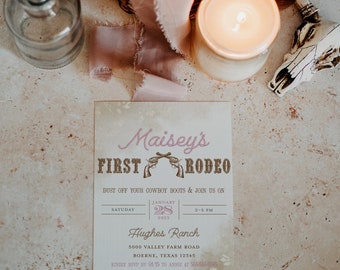 Könnte beinhalten: Eine rustikale Einladung zum ersten Rodeo eines Kindes. Die Einladung ist auf strukturiertem Papier mit einem rosa-goldenen Farbschema gedruckt. Der Text lautet "Maisey's First Rodeo" mit einem Paar rosa Cowboy-Stiefeln und dem Datum "January 28, 2023". Die Einladung enthält auch den Ort, "Hughes Ranch", und die Zeit, "2-5 PM".