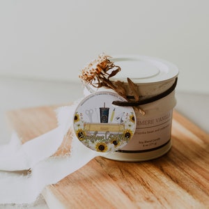 May include: A white candle jar with a brown ribbon and a round tag. The tag has a sunflower design and the text "Sunshine Creations" and "Personalized Tumblers, Shirts, and More". The candle jar label reads "Cashmere Vanilla Soy Blend Candle 6 oz (171 g)" and "of tonka bean and cedarwood."