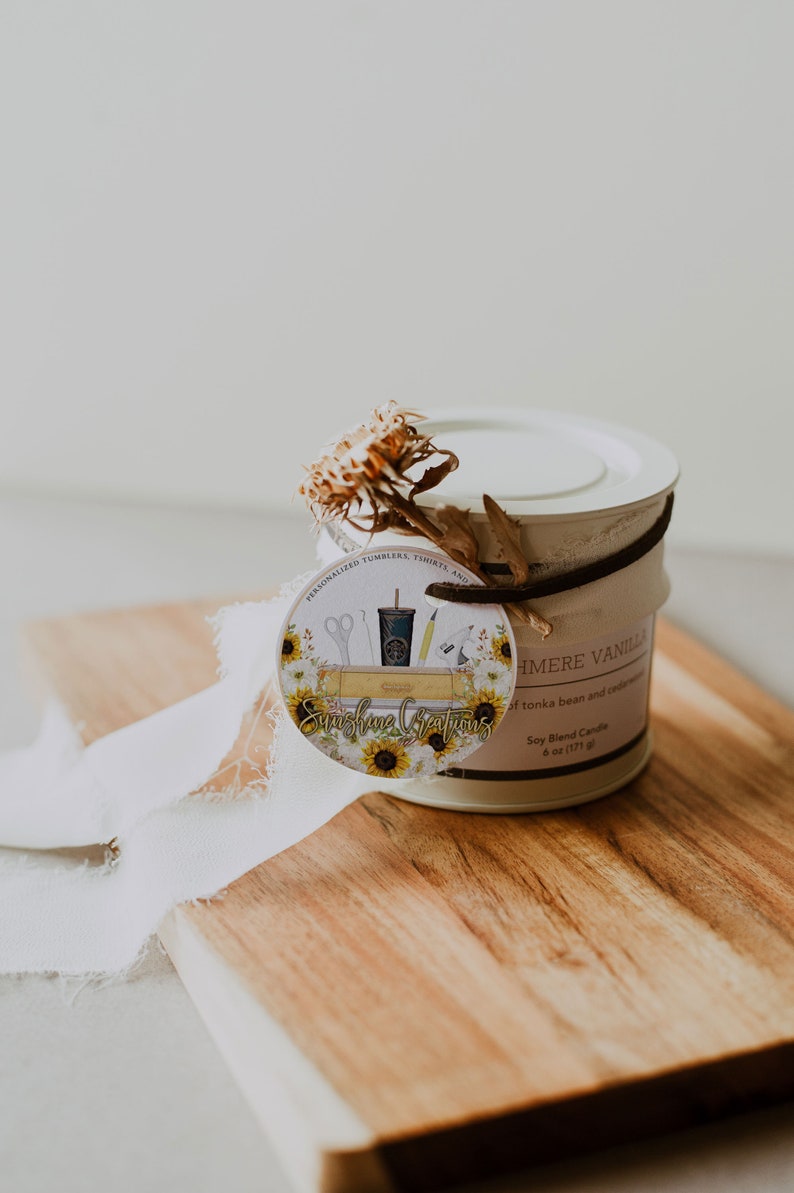 May include: A white candle jar with a brown ribbon and a round tag. The tag has a sunflower design and the text "Sunshine Creations" and "Personalized Tumblers, Shirts, and More". The candle jar label reads "Cashmere Vanilla Soy Blend Candle 6 oz (171 g)" and "of tonka bean and cedarwood."
