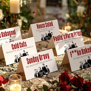 Könnte beinhalten: Weiße Tischkarten mit Menüpunkten wie "The Gabagool" und "Sunday Gravy" sind auf einem Tisch ausgestellt. Jede Karte zeigt ein Schwarz-Weiß-Bild einer Männergruppe. Rote Rosen und brennende Kerzen umgeben die Karten und schaffen eine festliche Atmosphäre.