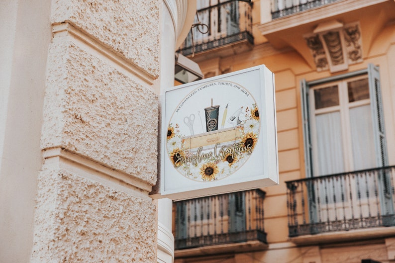 May include: A white sign with a round logo featuring sunflowers and the text "Sunshine Creations" with the tagline "Personalized Tumblers, Totes, and More".