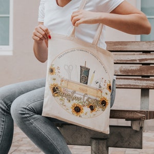 May include: A white canvas tote bag with a round design featuring sunflowers, crafting tools, and the text "Sunshine Creations" in a yellow script font. The text above the design reads "Personalized Tumblers, Shirts, and More".