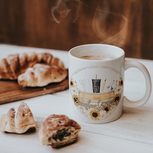 May include: White ceramic mug with a sunflower design and the text "Sunshine Crafts" on a white wooden table with croissants.