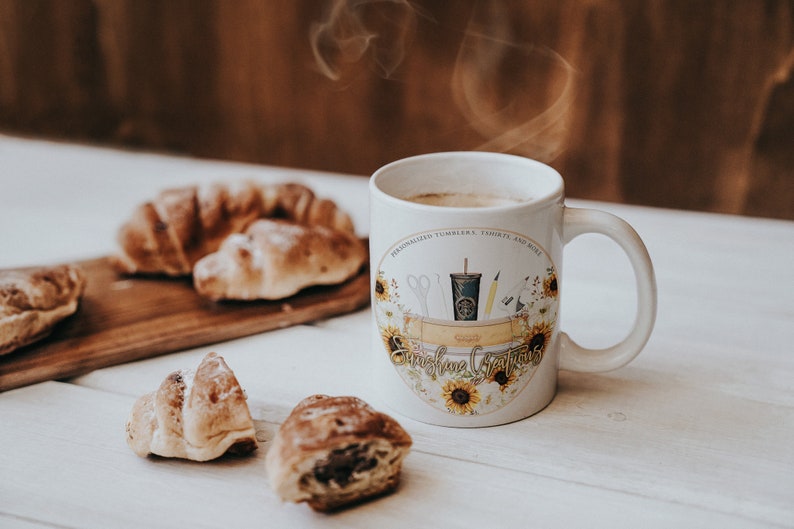 May include: White ceramic mug with a sunflower design and the text "Sunshine Crafts" on a white wooden table with croissants.