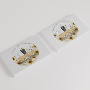 May include: Two white business cards with a circular logo featuring a sunflower design. The logo includes a wooden shelf with a cup of coffee, scissors, a pencil, and a ruler. The text "Sunshine Creations" is written in a cursive font.