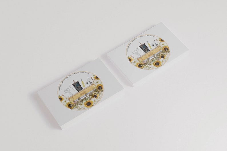 May include: Two white business cards with a circular logo featuring a sunflower design. The logo includes a wooden shelf with a cup of coffee, scissors, a pencil, and a ruler. The text "Sunshine Creations" is written in a cursive font.