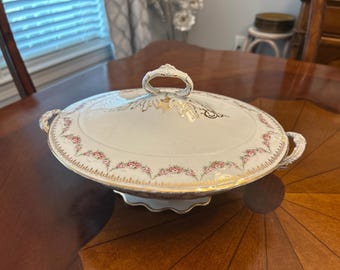 May include: An oval, lidded serving dish with a white base, adorned with pink floral patterns and gold trim. The dish has a decorative handle on the lid and two side handles. The dish is on a wooden table.