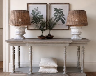 May include: A long wooden console table with turned legs and a lower shelf holding two white pillows. Two lamps with white weathered urn bases and brown woven wicker shades sit on top, flanking two framed botanical prints and two small potted plants.
