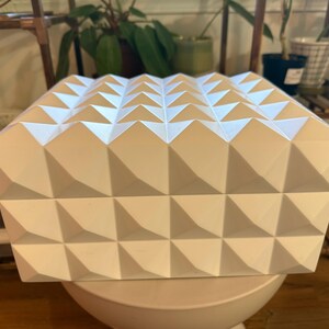 May include: A white, geometric box with a repeating triangular pattern on the top and sides. The box is sitting on a white, rounded base. The design creates a textured, three-dimensional effect. The box is likely a decorative storage container or a unique gift box.