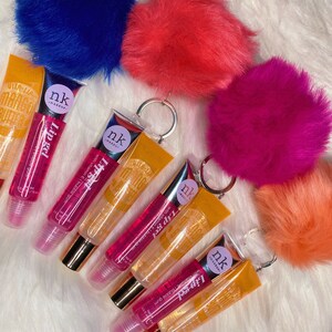 May include: Five clear lip gloss tubes with gold caps and colorful pom-pom keychains. The lip glosses are labeled "nk makeup" and "Lip gel" with a mango design. The keychains are blue, pink, red, and orange.