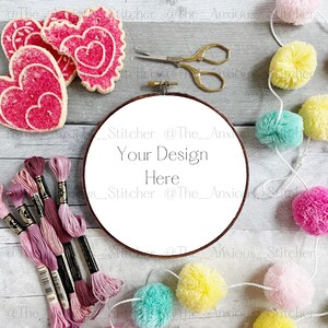 Op de afbeelding: Een witte borduurring met de tekst "_Stitcher @The_Anx Your Design Here _Stitcher @The_Anxious" op een grijze houten achtergrond.  Roze, gele en blauwe pompoenen zijn aan een wit touw rond de ring geregen.  Roze hartvormige koekjes en roze borduurdraad liggen ook op de achtergrond.