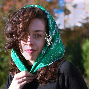 May include: A woman with curly brown hair wears a green headscarf with a white floral pattern. The scarf is tied at the neck and the ends are tucked under her chin. She is wearing a black shirt.