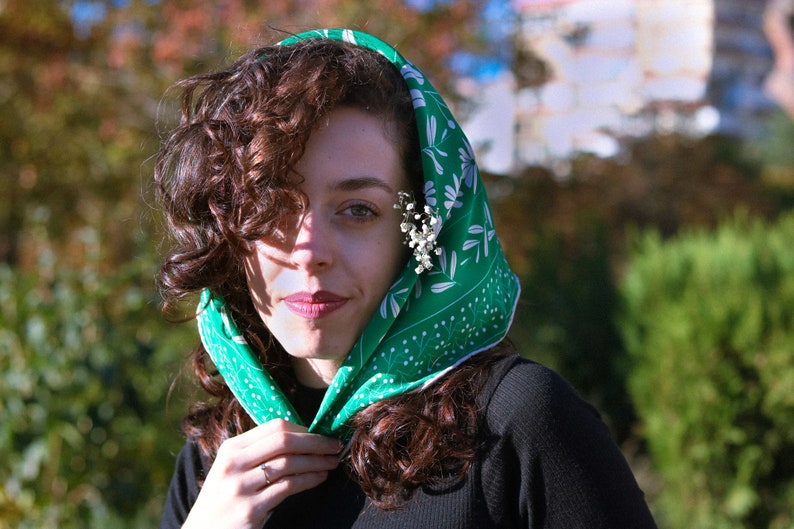 May include: A woman with curly brown hair wears a green headscarf with a white floral pattern. The scarf is tied at the neck and the ends are tucked under her chin. She is wearing a black shirt.