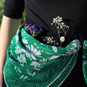 May include: A green floral patterned scarf tied around the waist of a person wearing a black shirt. The scarf has white flowers and green leaves on a green background. The scarf is tied in a knot at the front and the ends are hanging down.