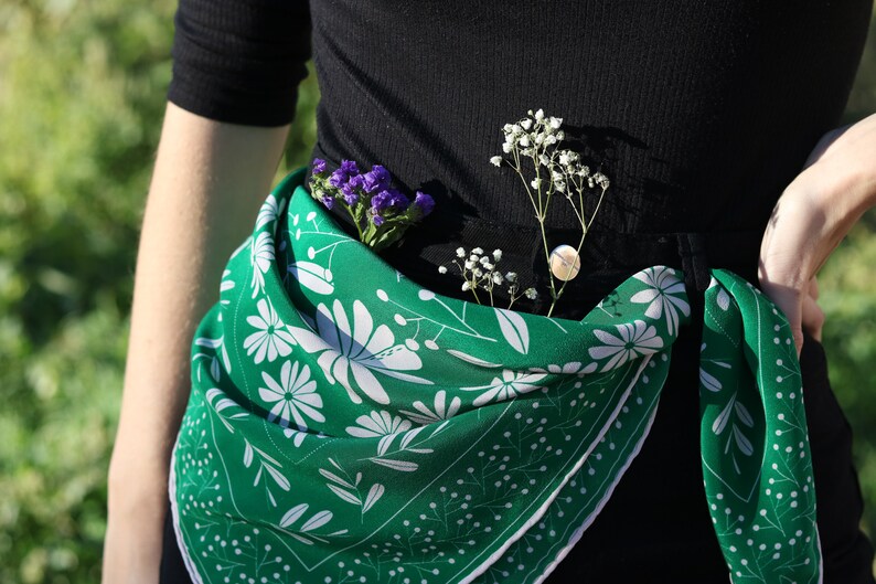 May include: A green floral patterned scarf tied around the waist of a person wearing a black shirt. The scarf has white flowers and green leaves on a green background. The scarf is tied in a knot at the front and the ends are hanging down.