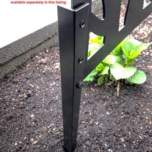 a close-up view of a black metal ground stake attached to a metal frame, with a small green plant growing next to it.