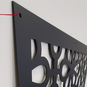 May include: A black decorative wall panel with a geometric cut-out design. The panel is mounted on a white wall and has a matte finish. The panel has a series of holes for mounting.