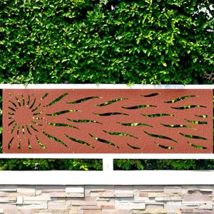 May include: A rectangular, rust-colored metal fence panel with a sun and wave design. The sun is on the left, and the waves extend to the right. The panel is mounted on a white fence, with green foliage in the background.