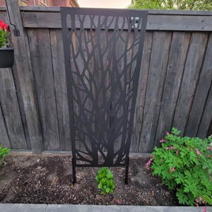 a black metal trellis with a tree-like design, situated in a garden setting with a wooden fence and various plants.