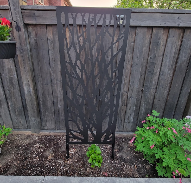 a black metal trellis with a tree-like design, situated in a garden setting with a wooden fence and various plants.
