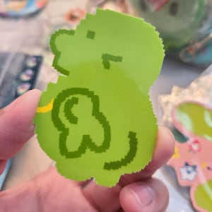 May include: A bright green, pixelated sticker in the shape of a cartoon character. The sticker has a rounded shape with a yellow accent and a dark green outline. Other stickers are visible in the background.