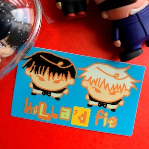 May include: A blue card with cartoon illustrations of two characters with black and white hair. The card has the text "Hello Fit" in orange and green. The background is red, and there are small figurines in the image.