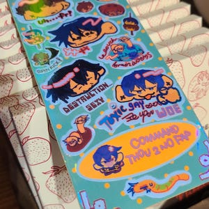 May include: A rectangular sticker sheet with a light blue background featuring various cartoon characters and text including "DESZJULU", "uh...fr?", "SPIN KICK!", "DESTRUCTION...SEXY", "TOXIC GAY", "brain worms", "COMMAND THOU 2 NO FAP", and "be upon WOE".