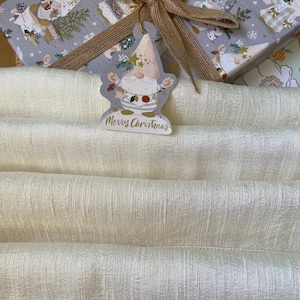 Puede incluir: Un envoltorio de regalo de tela blanca con una etiqueta de regalo con forma de gnomo que dice "Feliz Navidad". La etiqueta de regalo est&aacute; unida a una caja de regalo con estampado marr&oacute;n y blanco con una cinta marr&oacute;n.