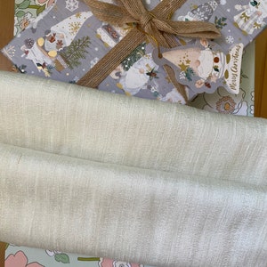 Puede incluir: Una caja de regalo gris con una cinta marr&oacute;n y un dise&ntilde;o de gnomo. La caja est&aacute; envuelta en papel que dice "Feliz Navidad". La caja est&aacute; sentada encima de una tela blanca y un papel de envolver con estampado floral.