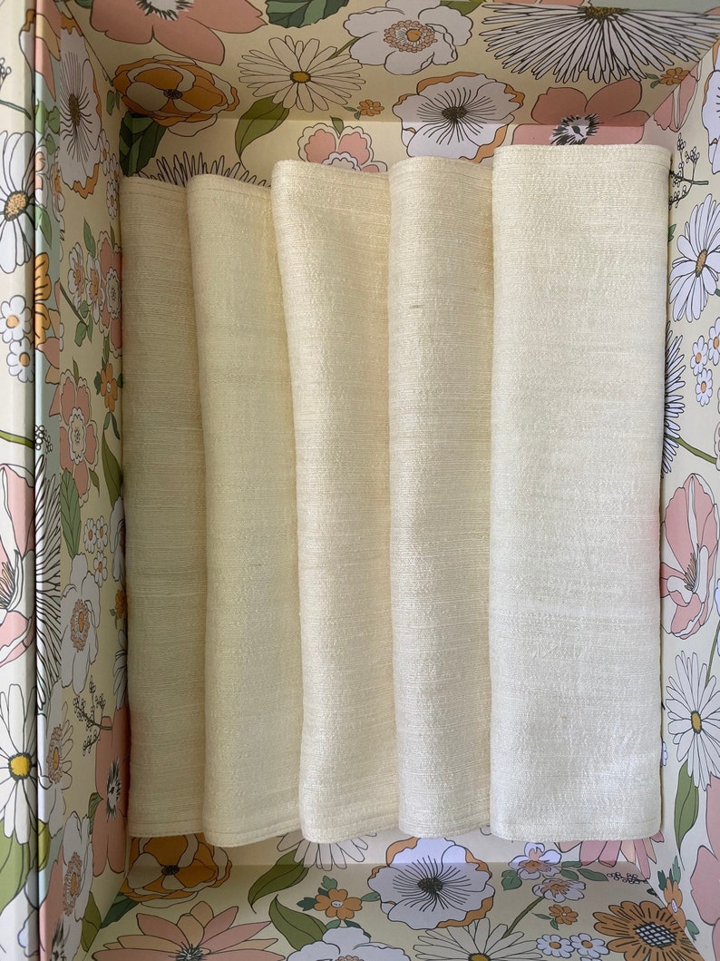 Puede incluir: Cinco servilletas de lino de color crema dobladas y apiladas dentro de una caja con estampado floral.